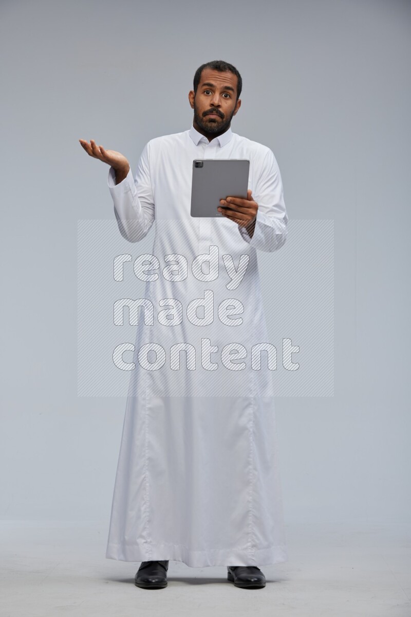 Saudi man wearing Thob standing working on tablet on Gray background