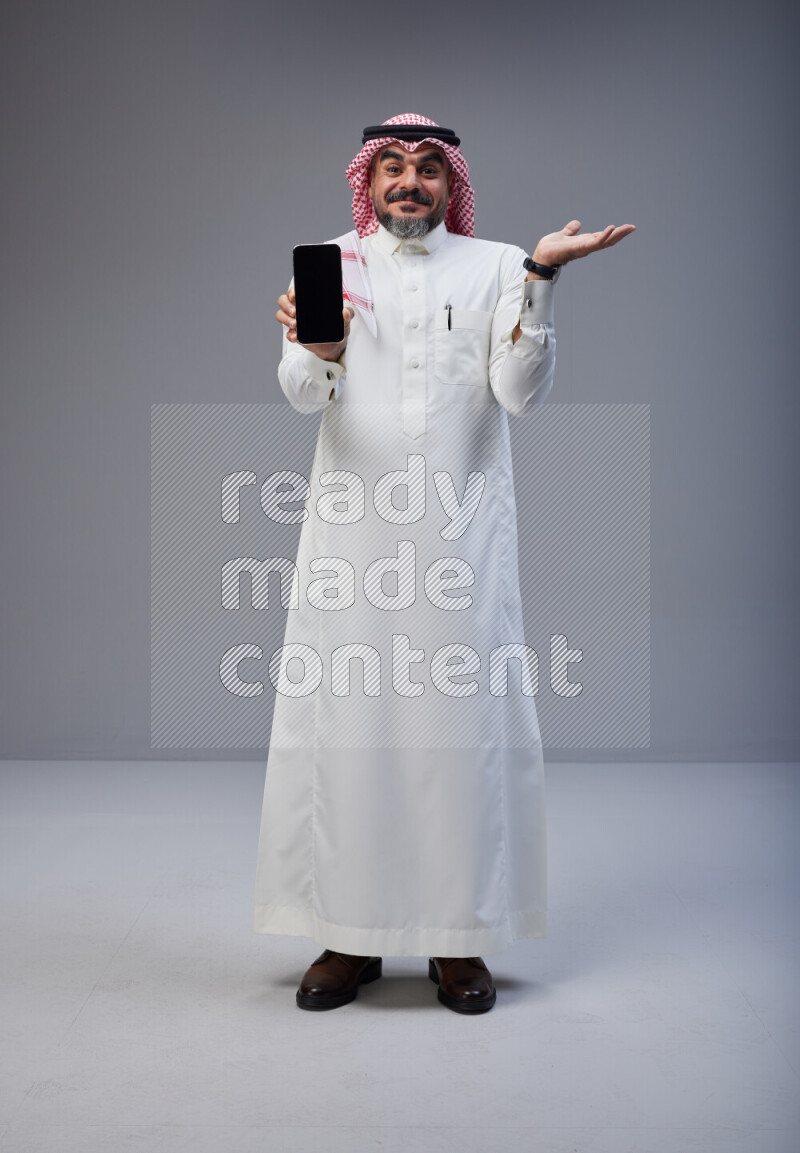 Saudi man Wearing Thob and red Shomag standing showing phone to camera on Gray background