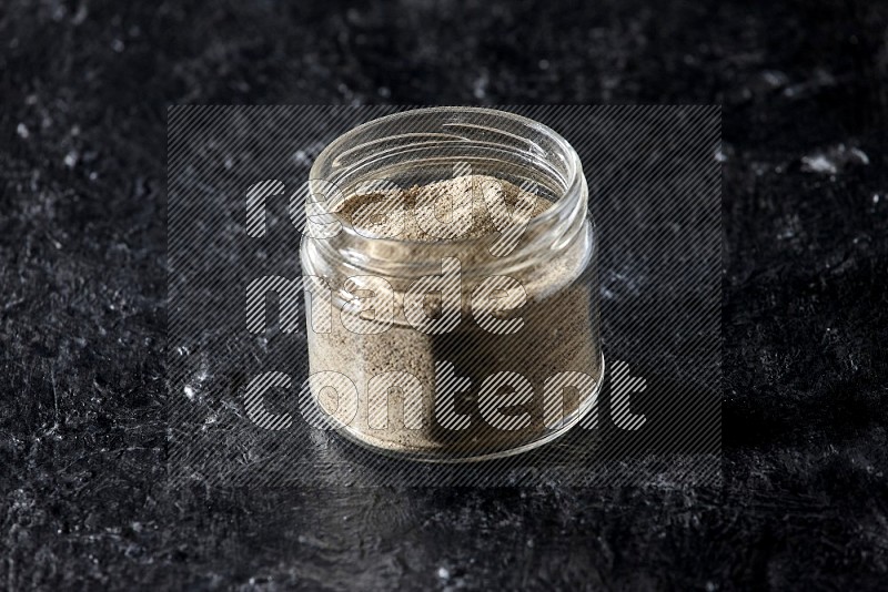 A glass jar full of white pepper powder on textured black flooring