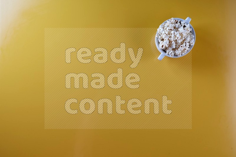 A white ceramic bowl full of popcorn on a yellow background in a top view shot