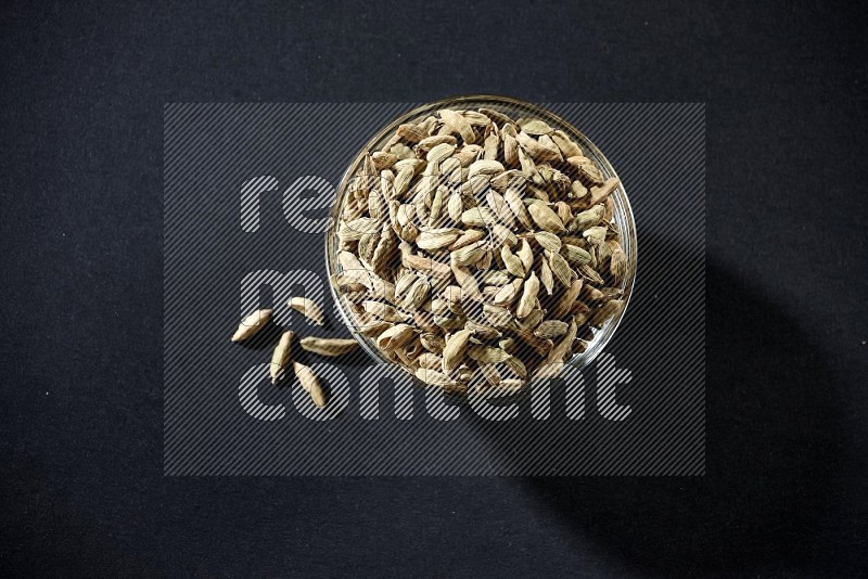 A glass bowl full of cardamom and more seeds spreaded beneath the bowl on black flooring