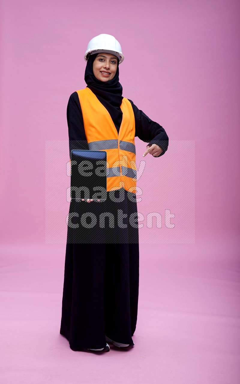 Saudi woman wearing Abaya with engineer vest and helmet standing showing tablet to camera on pink background