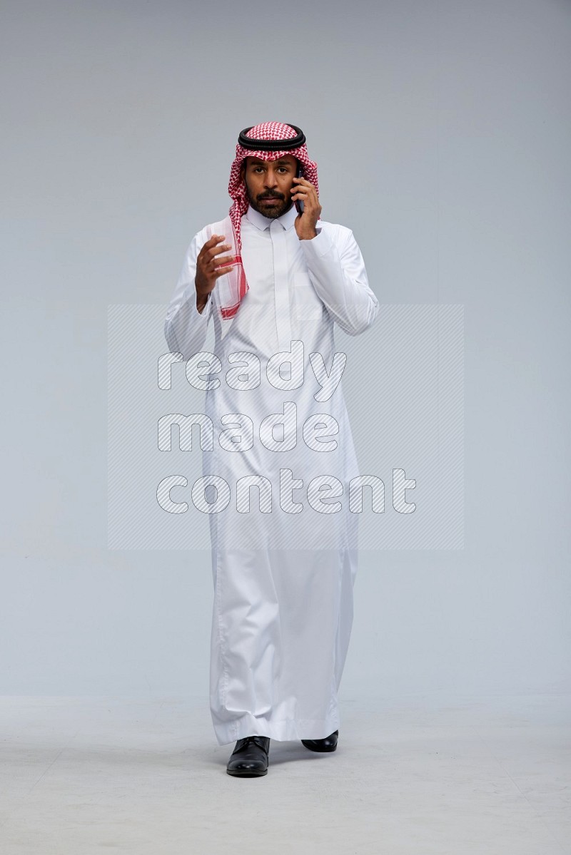 Saudi man Wearing Thob and shomag standing talking on phone on Gray background