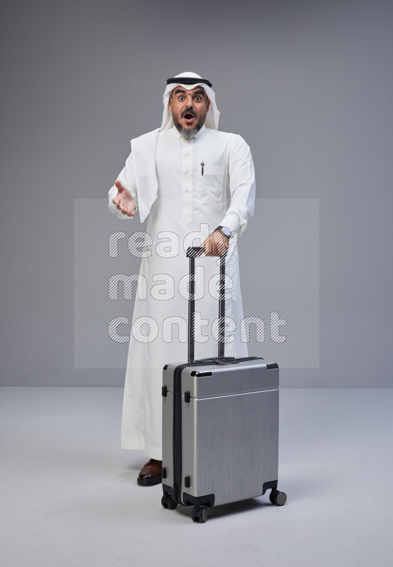 Saudi man wearing Thob and white Shomag standing holding Travel bag on Gray background