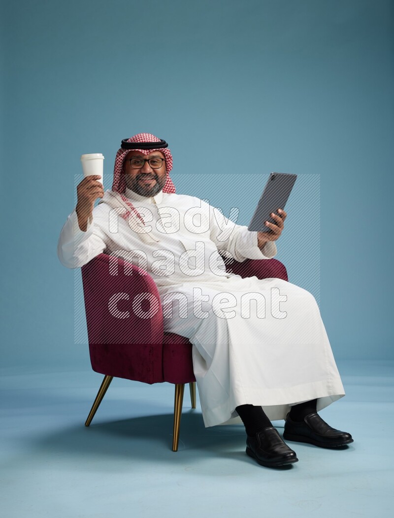 Saudi Man with shimag sitting on chair working on tablet on blue background