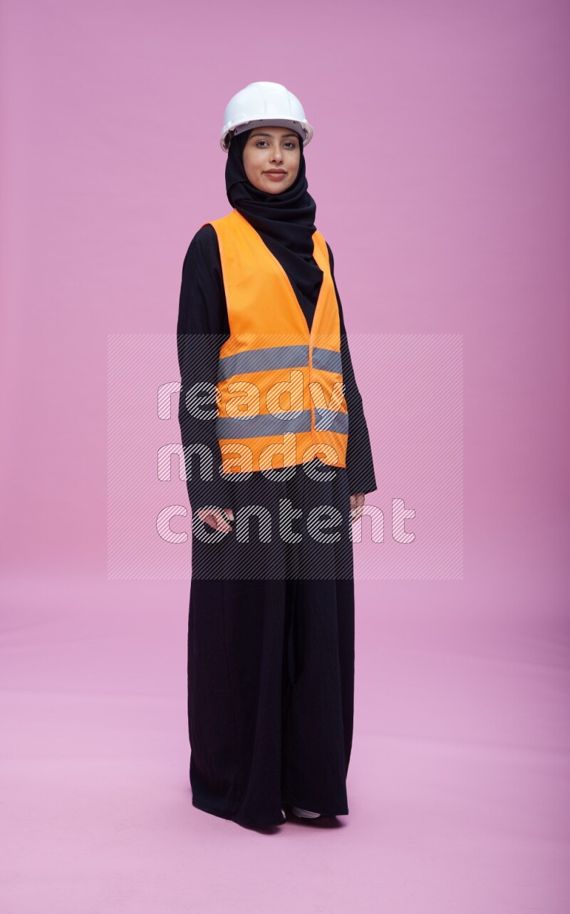 Saudi woman wearing Abaya with engineer vest and helmet standing interacting with the camera on pink background