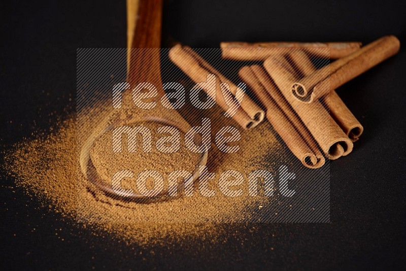 Cinnamon powder in a wooden ladle spoon with cinnamon sticks on black background
