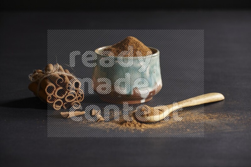 Ceramic bowl full of cinnamon powder and a wooden spoon full of powder with cinnamon sticks stacked and bounded on black background