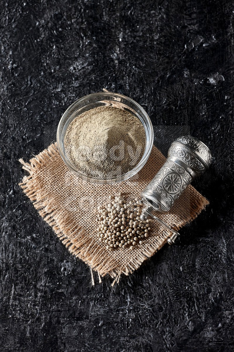 A glass bowl full of white pepper powder with white pepper beads on a burlap piece of fabric and a metal grinder on textured black flooring