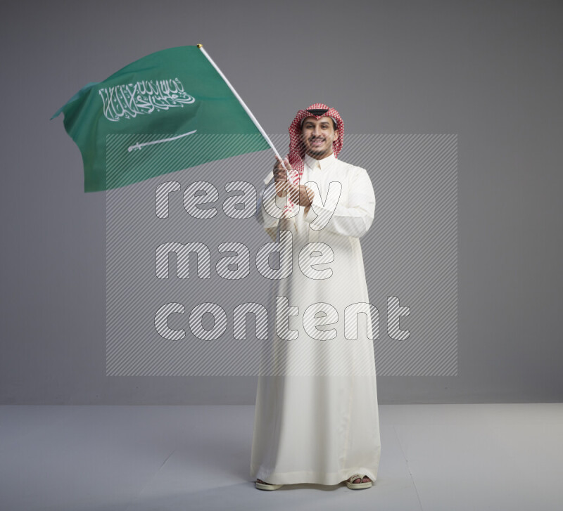 A Saudi man standing wearing thob and red shomag raising big Saudi flag on gray background