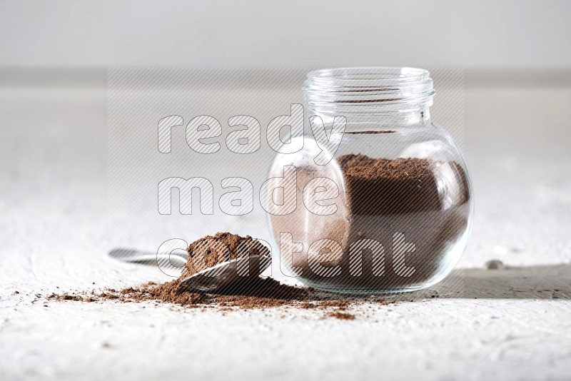 A glass spice jar and a metal spoon full of cloves powder on textured white flooring