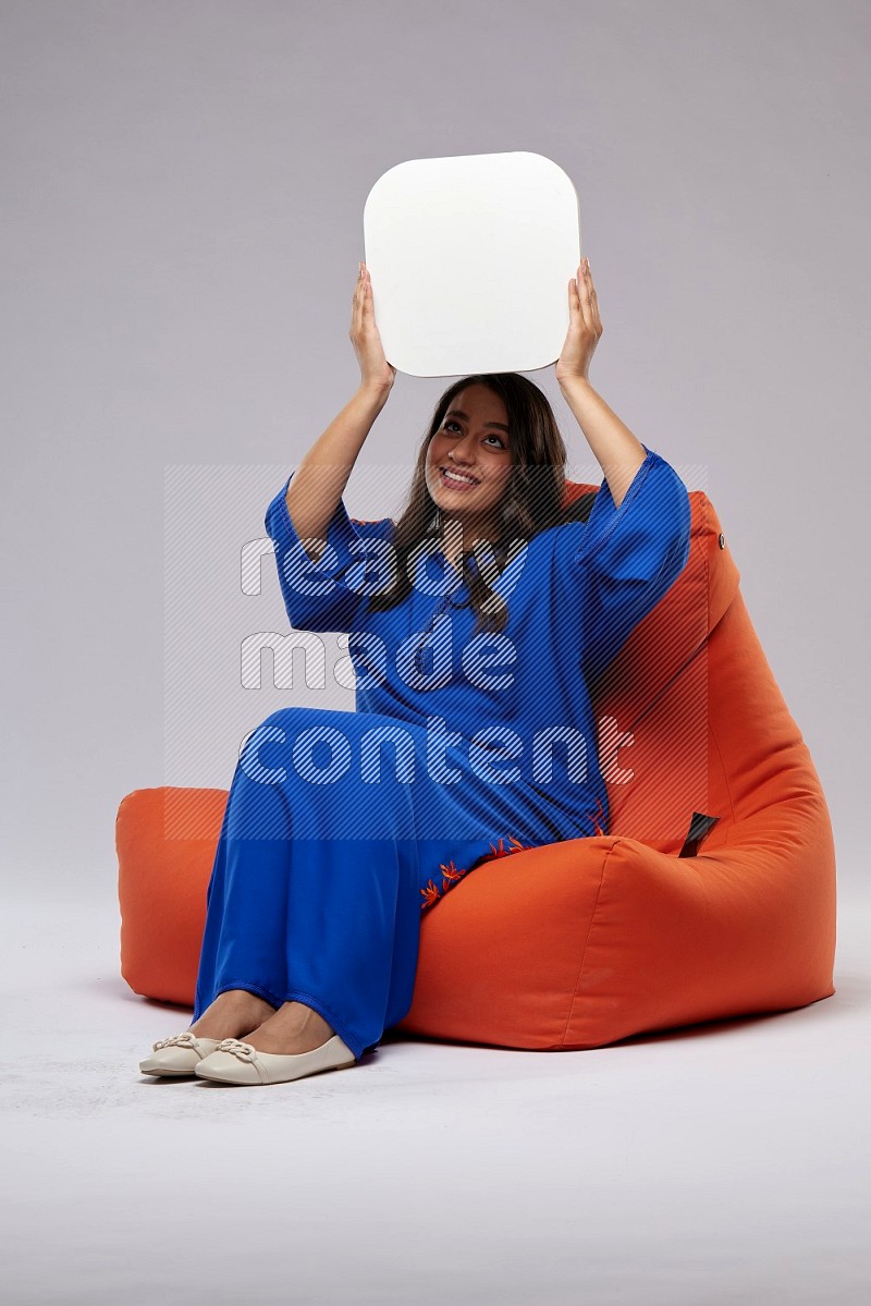 A Woman sitting on an orange beanbag wearing Jalabeya holding a social media sign