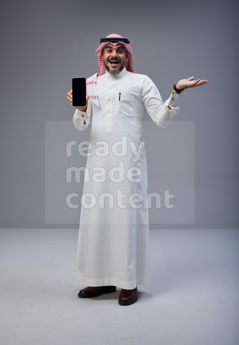Saudi man Wearing Thob and red Shomag standing showing phone to camera on Gray background