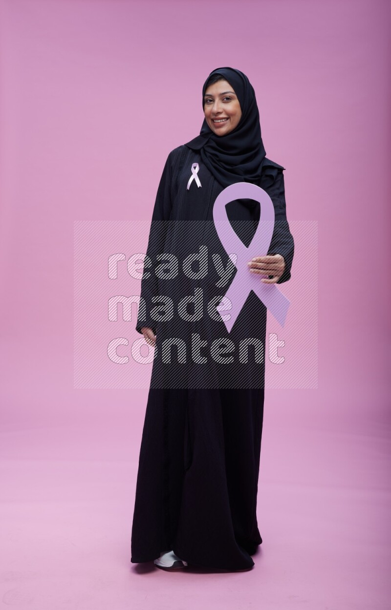 Saudi woman wearing pink ribbon on abaya standing holding awareness ribbon on plain pink background