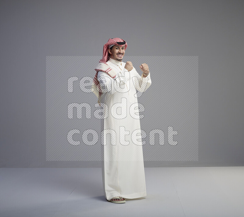 A Saudi man standing wearing thob and red shomag interacting with the camera on gray background