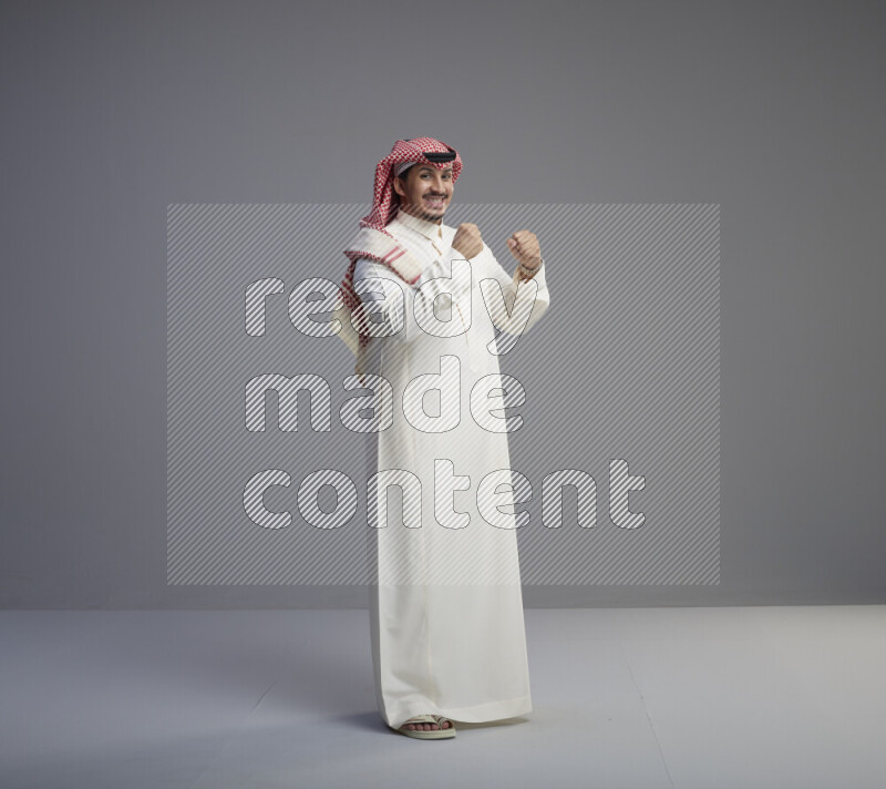 A Saudi man standing wearing thob and red shomag interacting with the camera on gray background