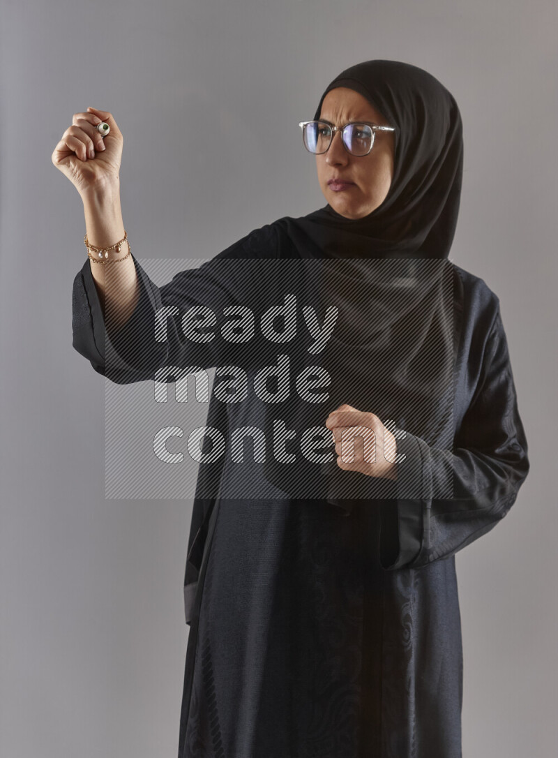 Woman in black abaya, hijab and glasses holding a marker pen to write on a transparent board with different reactions and expressions on grey background