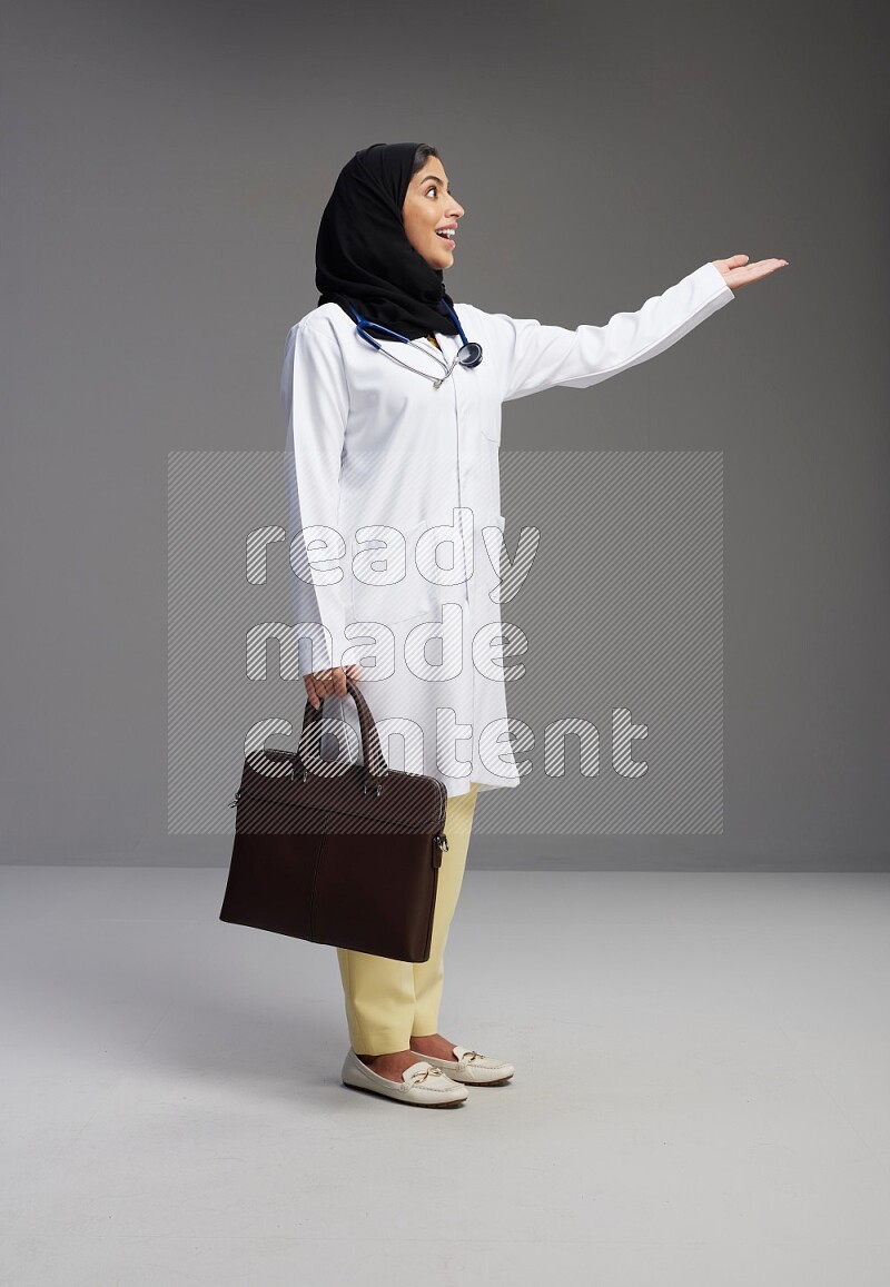 Saudi woman wearing lab coat with stethoscope standing holding bag on Gray background