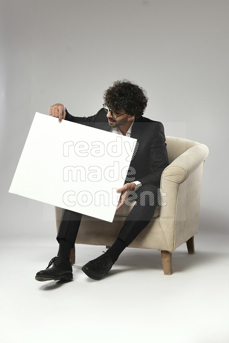 رجل يرتدي ملابس رسمية يجلس علي كرسي و يحمل لوحة بيضاء علي خلفية بيضاء