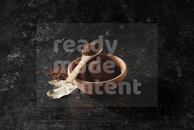 A wooden bowl and a wooden spoon full of cloves powder with laurel leaves on a textured black flooring