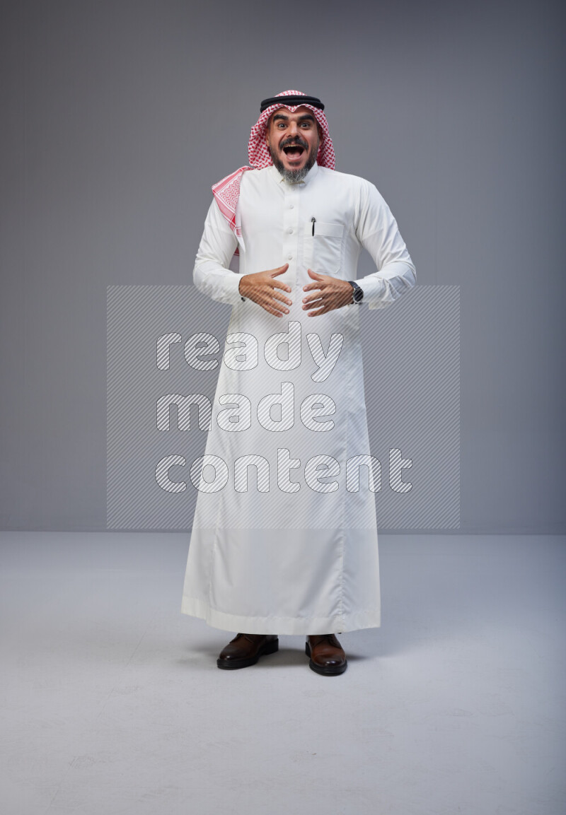 Saudi man Wearing Thob and red white Shomag standing interacting with the camera on Gray background