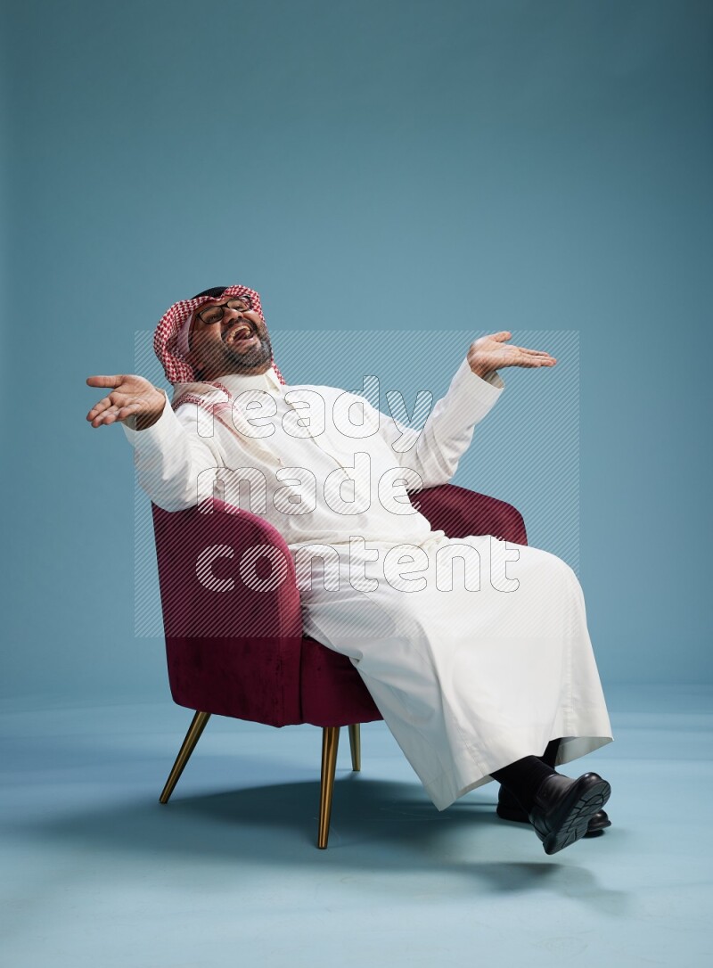 Saudi Man with shimag sitting on chair Interacting with the camera on blue background