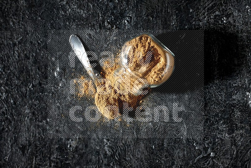 A flipped glass spice jar and a metal spoon full of allspice powder and powder spilled out of it on a textured black flooring