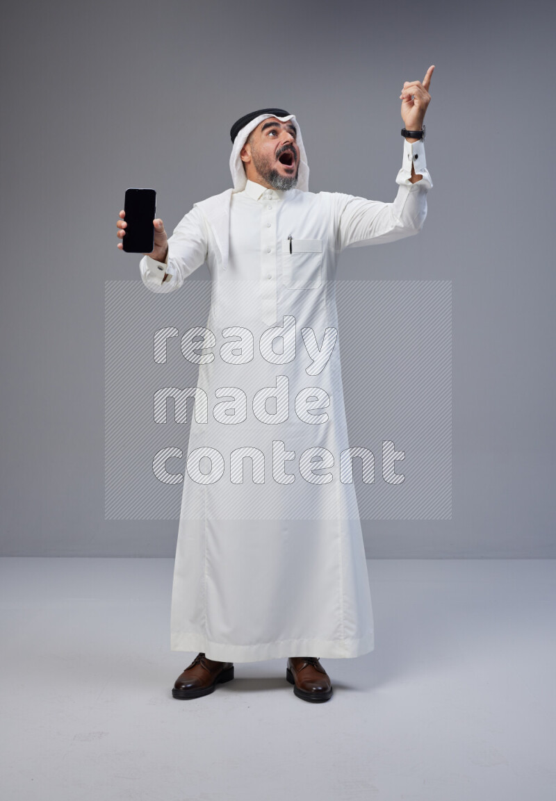 Saudi man Wearing Thob and white Shomag standing showing phone to camera on Gray background