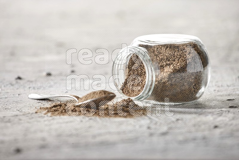Flipped glass spice jar full of black pepper powder with a metal spoon full of it on textured white flooring