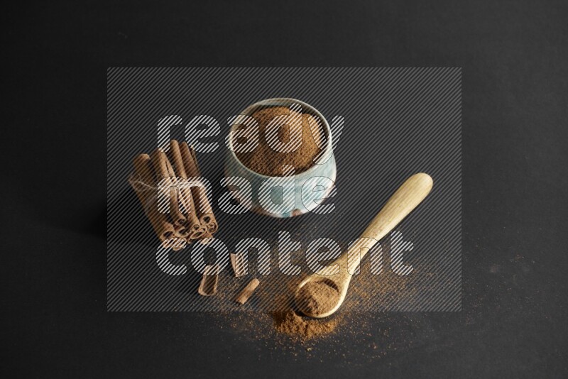 Ceramic bowl full of cinnamon powder and a wooden spoon full of powder with cinnamon sticks stacked and bounded on black background