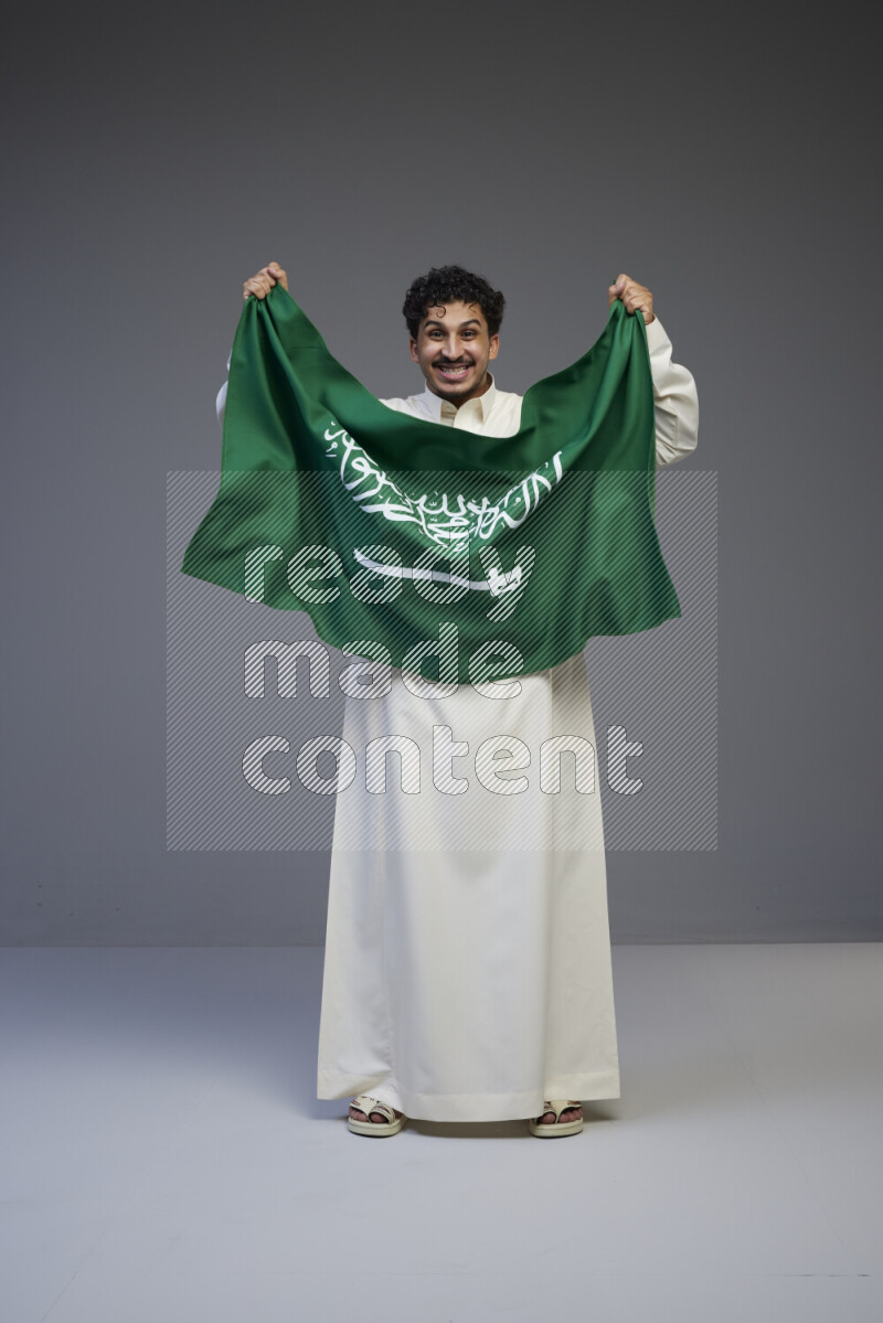 A Saudi man standing wearing thob holding big Saudi flag on gray background