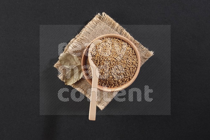 A wooden bowl and spoon full of mustard seeds on a piece of burlap on a black flooring