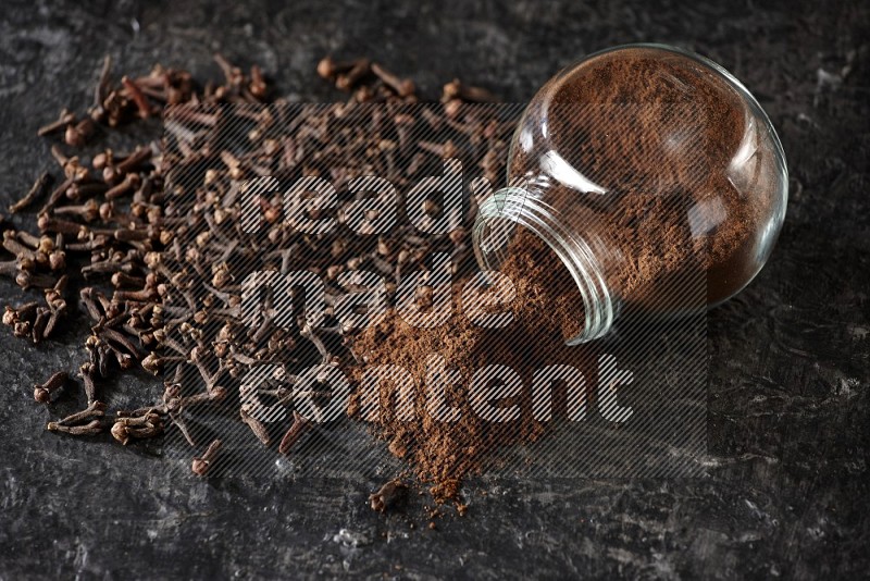 A flipped glass spice jar full of cloves powder and powder came out of it with cloves spread on textured black flooring