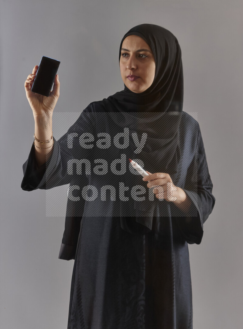 Woman in black abaya and hijab holding a marker pen to write on a transparent board with different reactions and expressions on grey background