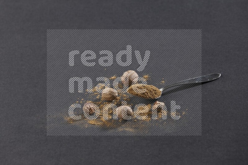 ملعقة معدنية ممتلئة ببودرة جوزة الطيب مع بودرة وحبوب جوزة الطيب بجانبها على خلفية سوداء