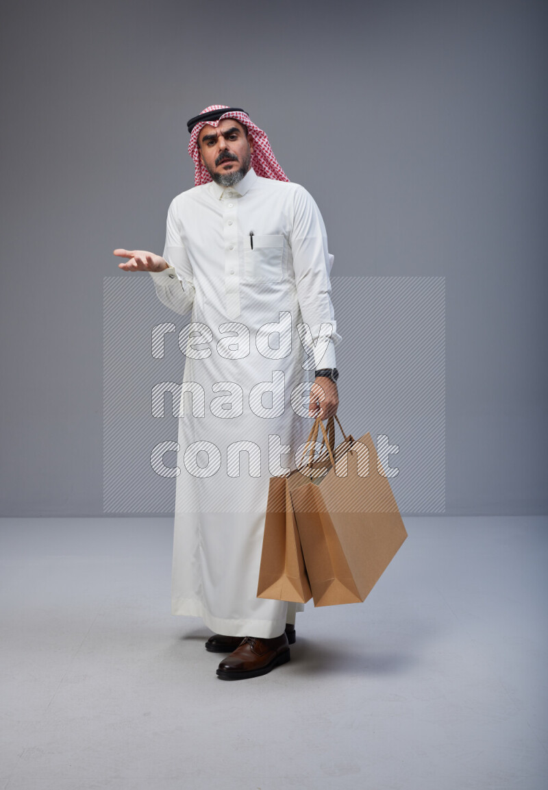 Saudi man Wearing Thob and red Shomag standing holding shopping bag on Gray background
