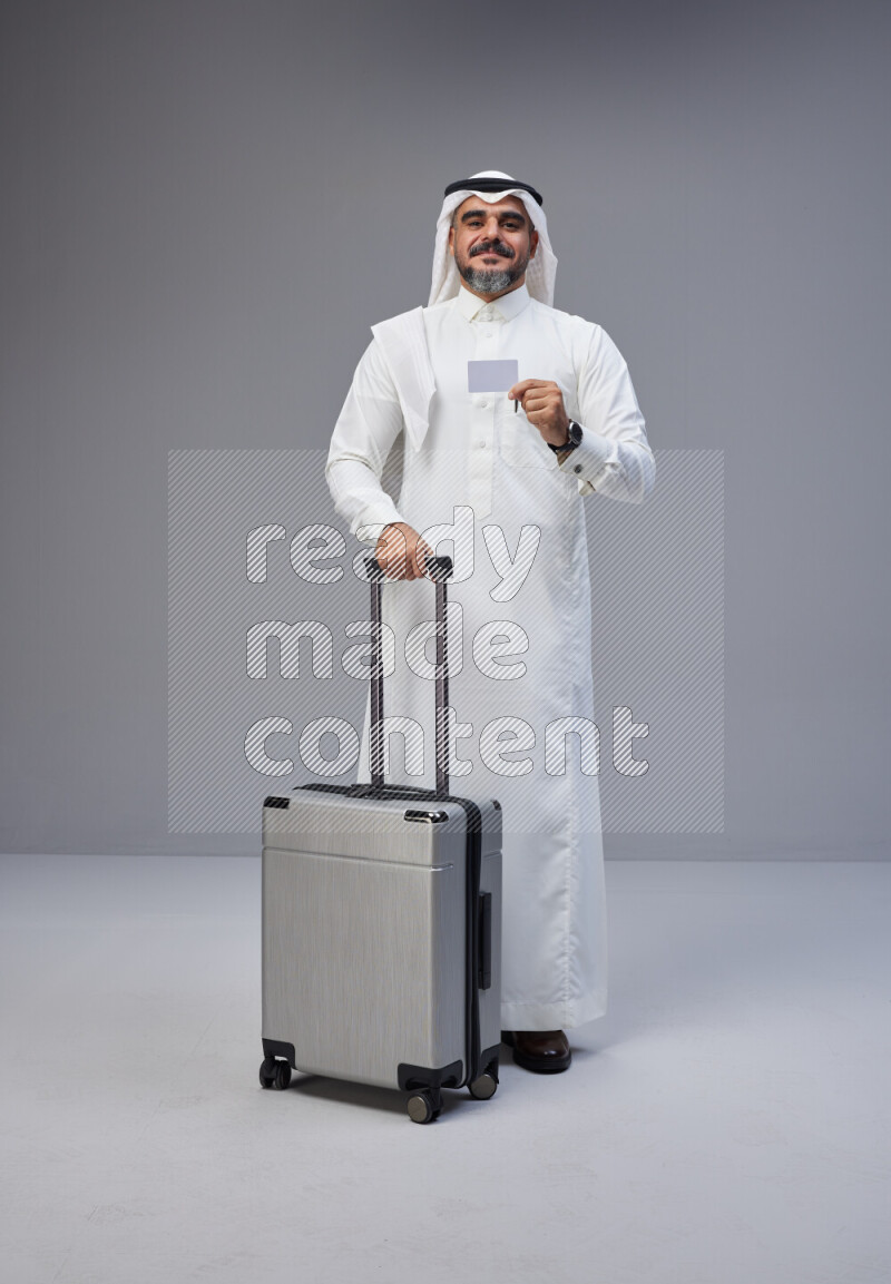 Saudi man wearing Thob and white Shomag standing holding Travel bag and ATM card on Gray background