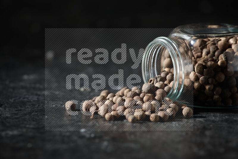 وعاء زجاجي للتوابل مقلوب ممتلئ بحبوب بهار الفلفل الحلو والحبوب تناثرت منه على خلفية سوداء