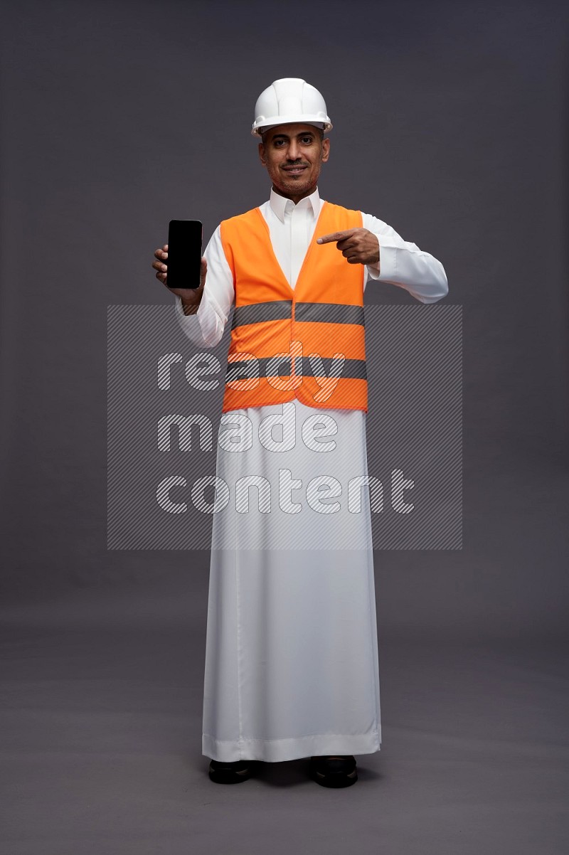 Saudi man wearing thob with engineer vest standing showing phone to camera on gray background