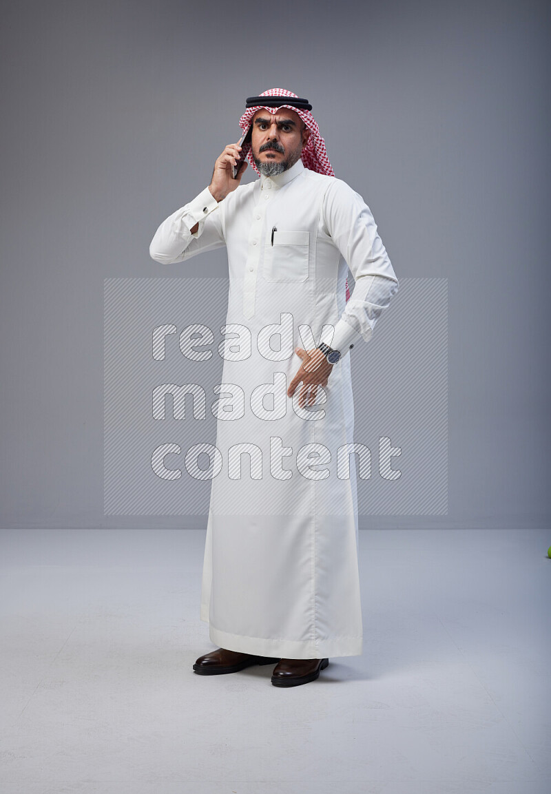 Saudi man Wearing Thob and red Shomag standing talking on phone on Gray background