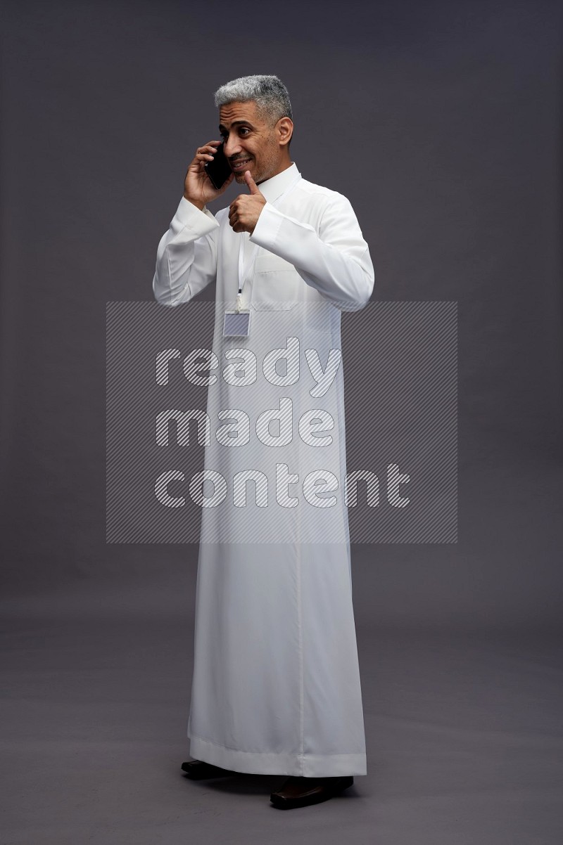 Saudi man wearing thob with neck strap employee badge standing talking on phone on gray background