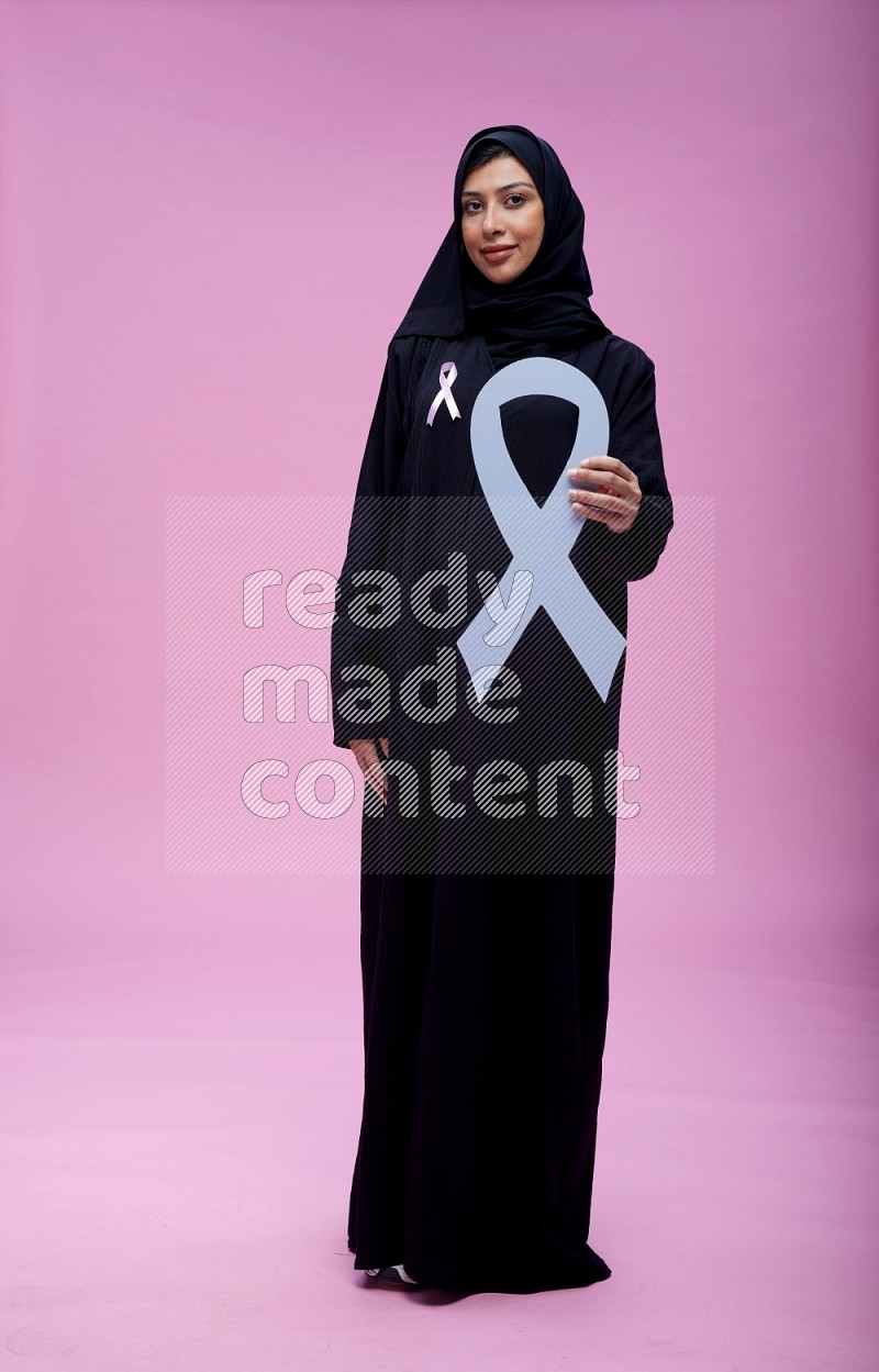 Saudi woman wearing pink ribbon on abaya standing holding awareness ribbon on plain pink background