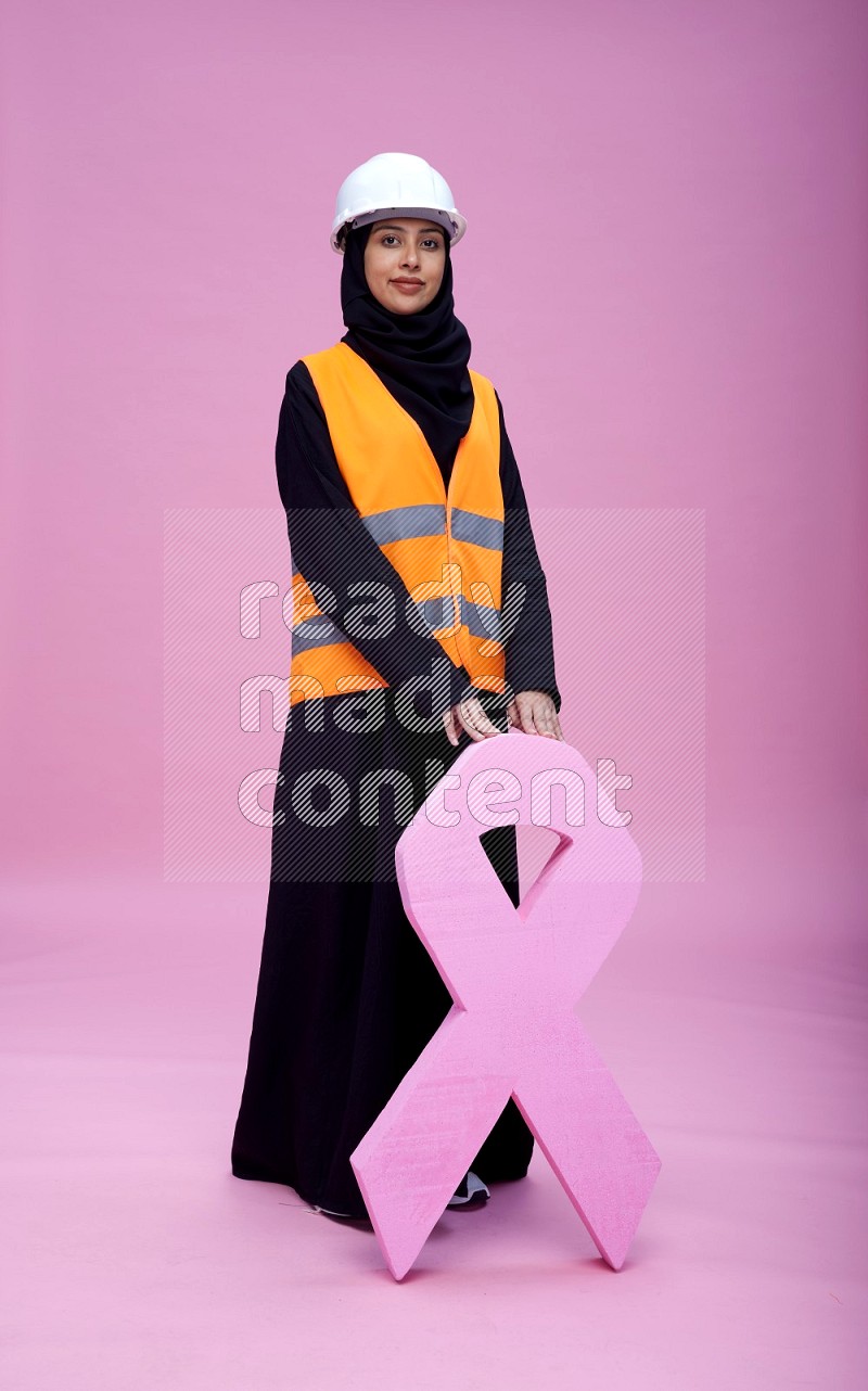 Saudi woman wearing Abaya with engineer vest and helmet standing awareness ribbon on pink background