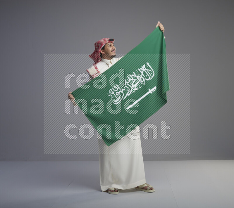 A saudi man standing wearing thob and red shomag holding big saudi flag on gray background