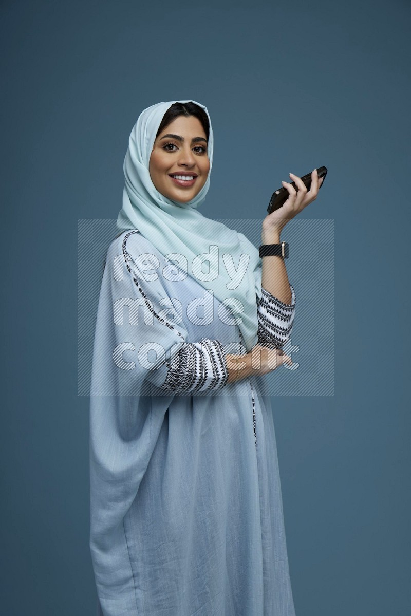 A Saudi woman posing with her phone in a blue background wearing a blue Abaya with hijab