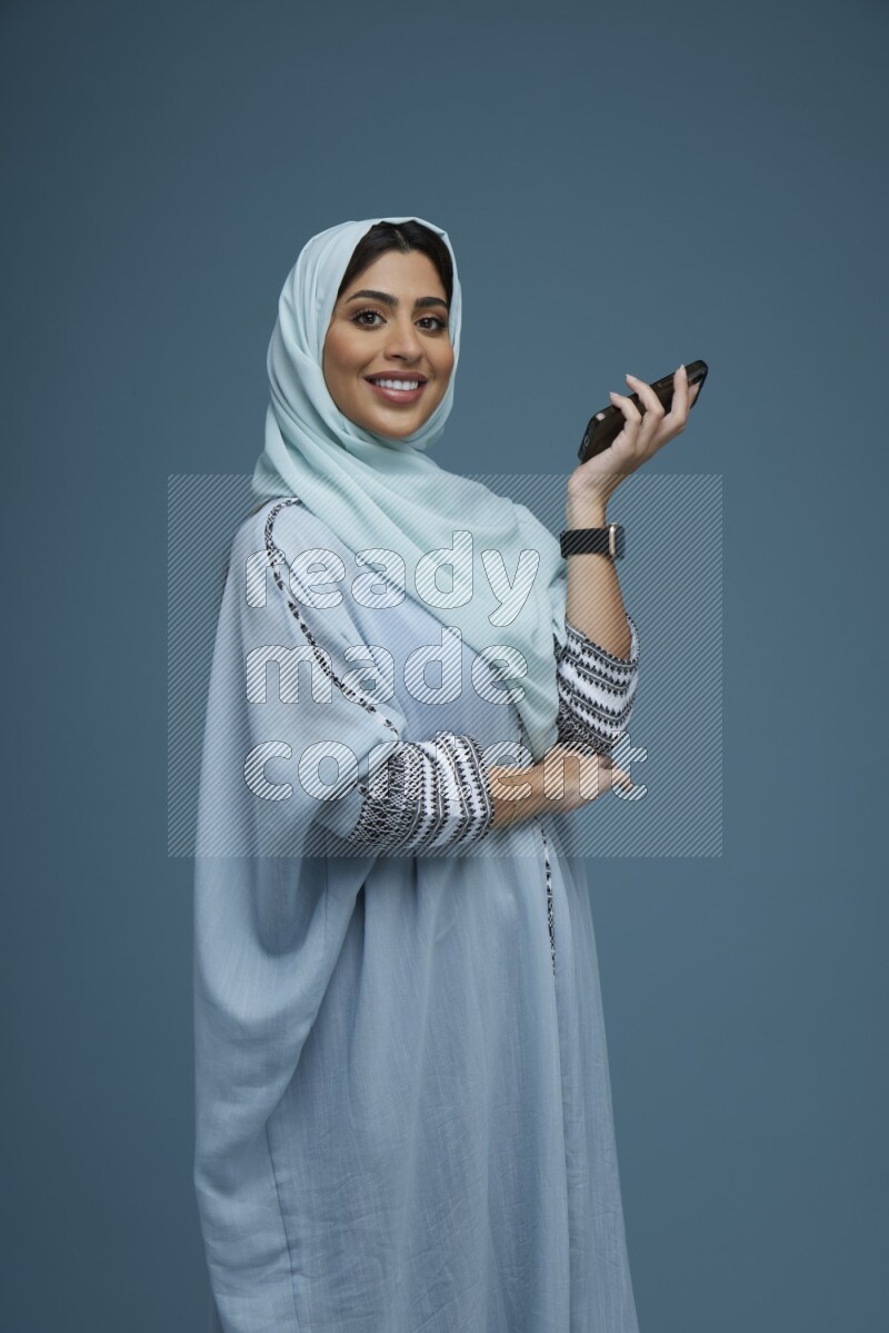 A Saudi woman posing with her phone in a blue background wearing a blue Abaya with hijab