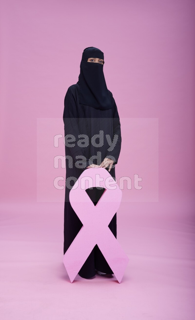 Saudi woman wearing abaya and niqab standing holding awareness ribbon on pink background