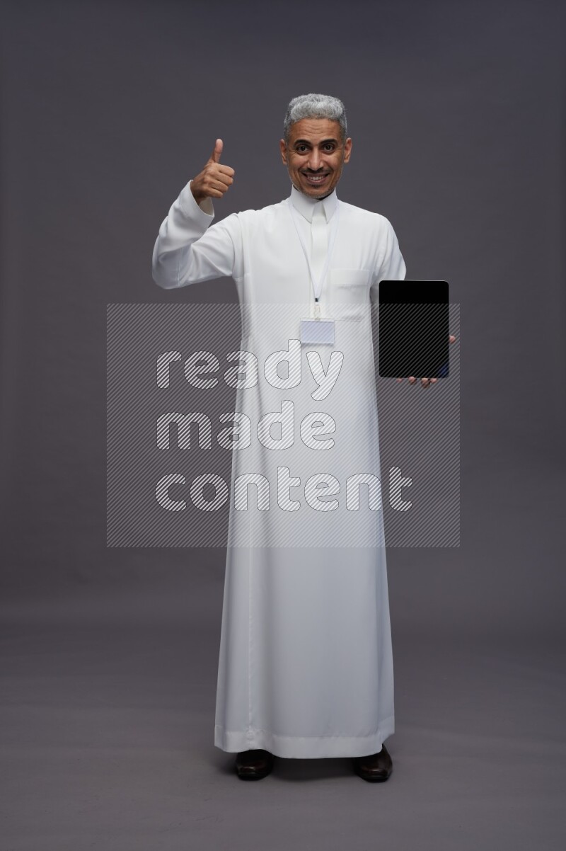 Saudi man wearing thob with neck strap employee badge standing showing tablet to camera on gray background