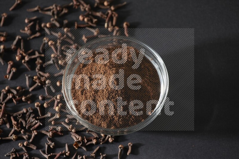 A glass bowl full of cloves powder with cloves grains spread on black flooring