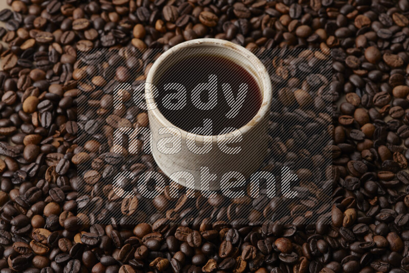A beige pottery cup of coffee surrounded by roasted coffee beans on beige background